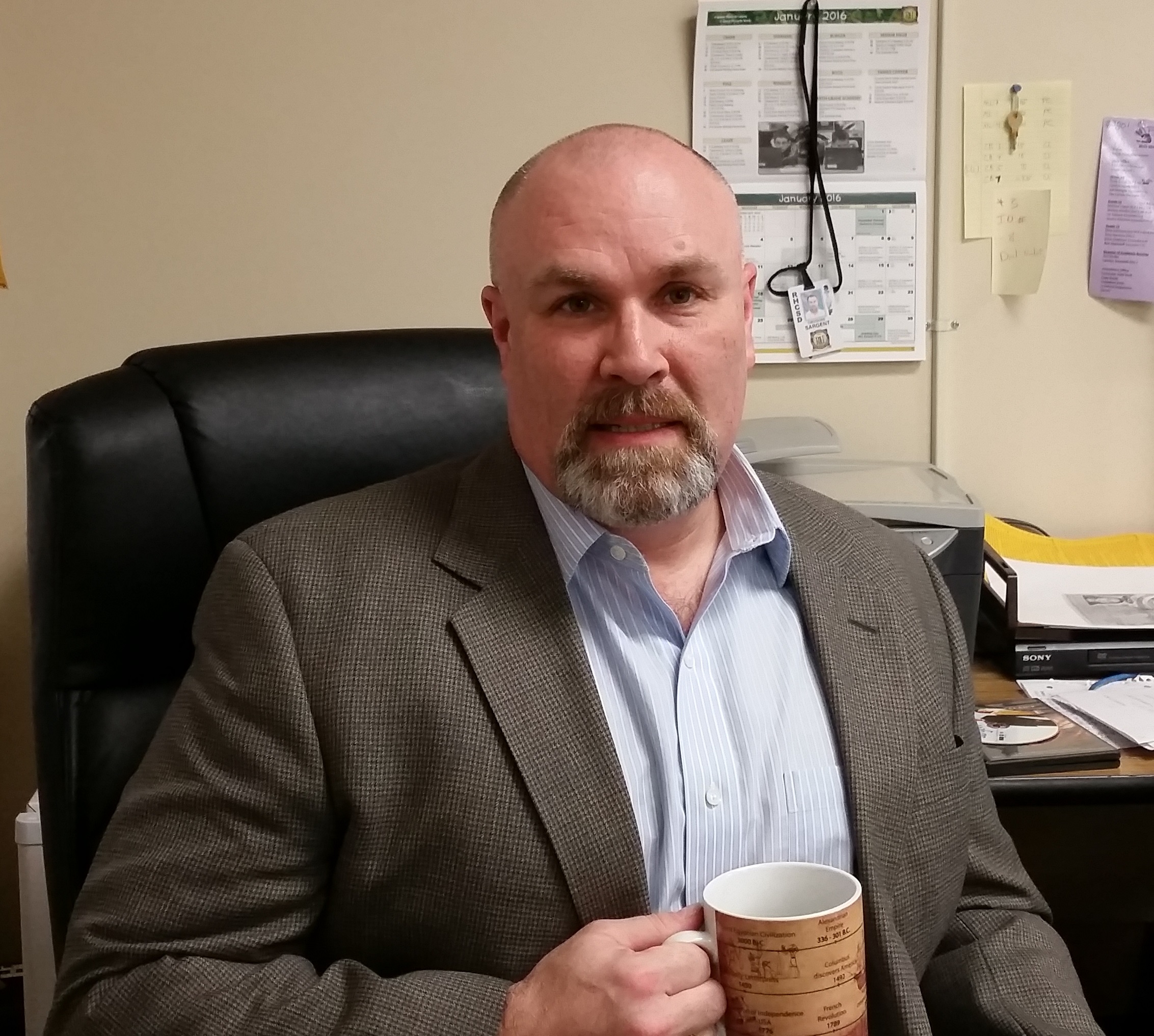 A man sits in a chair facing the camera. He has a goatee and is holding a coffee mug.