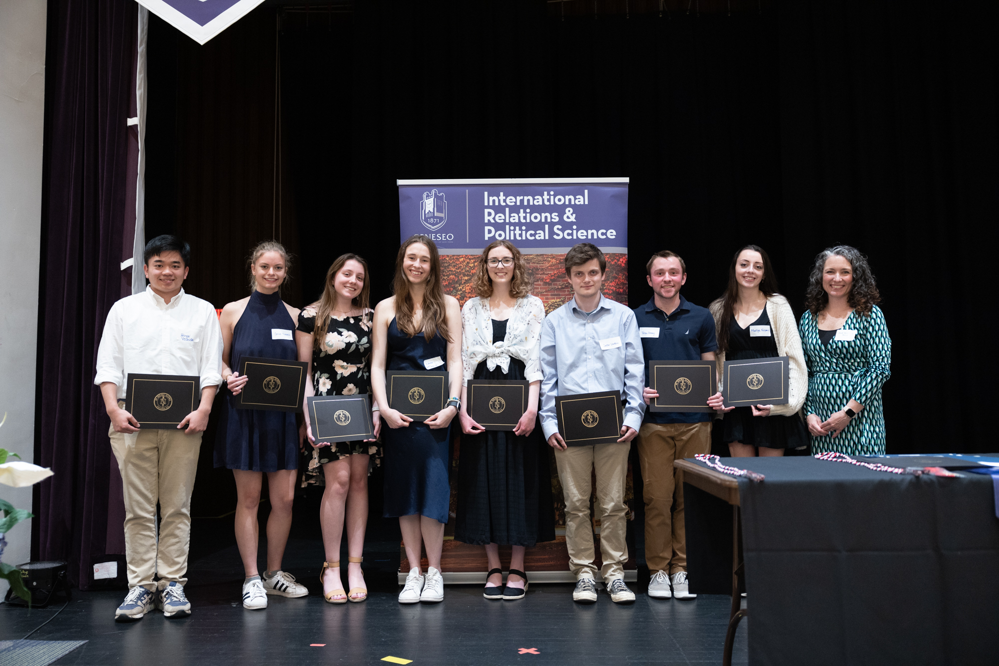 Eight students and a professor stand on a stage holding their awards certificates.