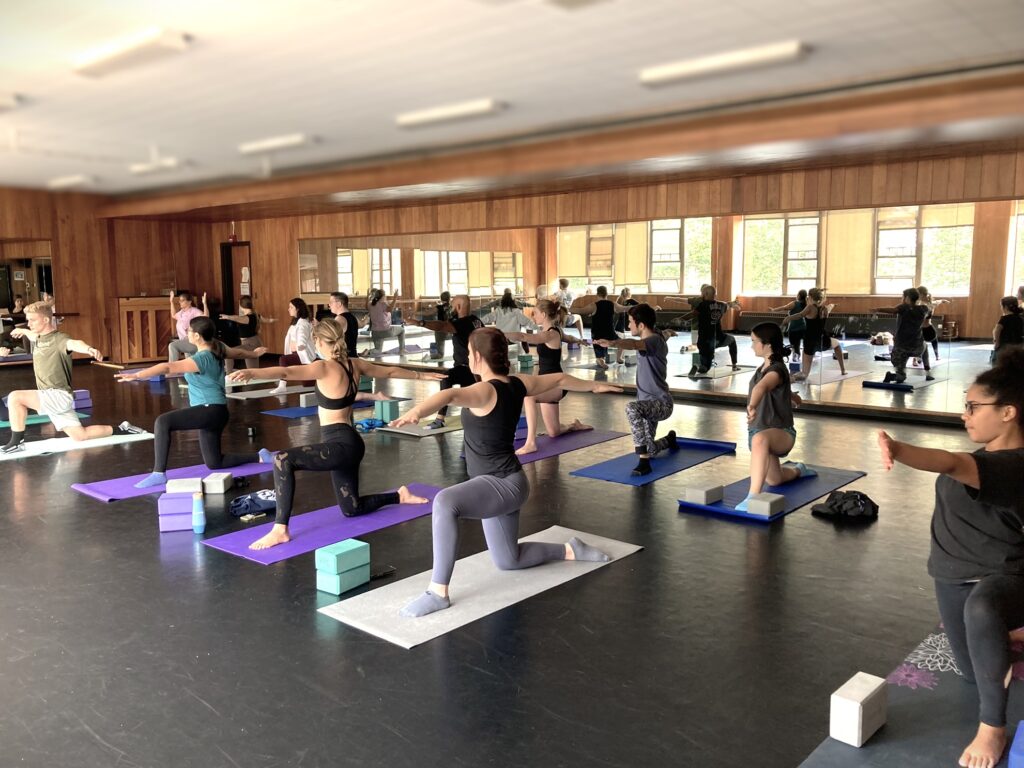 Approximately 12 students doing a yoga pose in a kneeling position on yoga mats in the Schrader Dance Studio, where this are mirrors in the background