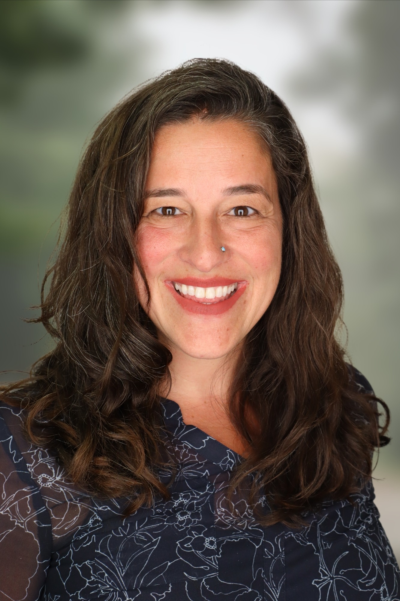 Feminine presenting person with shoulder length brown hair, smiles at the camera.