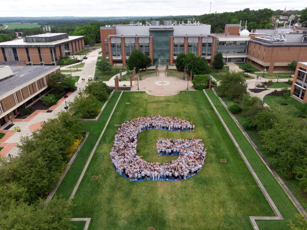 An aerial view of the SUNY Geneseo campus quad where a large group of students in grey shirts are standing together to form a giant letter "G" on the green lawn.