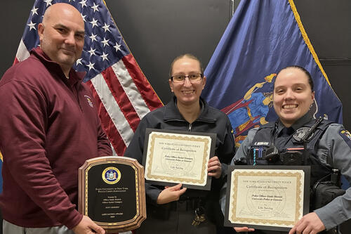 Three officers holding awards in front of an American flag and a NYS flag.