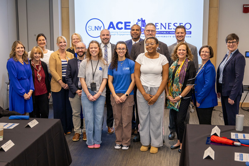 Group of about 15 people (including Assemblymember Andrea Bailey, members of the Geneseo College Council, and campus and SUNY leaders) posing for a photo in a conference room, standing in front of a SUNY ACE Geneseo presentation screen.